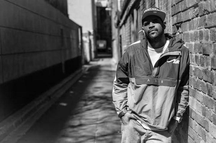 Jimblah is a Larrakia man with dark skin and a beard. He stands in a laneway, wearing a baseball cap and a Nike jacket, with his hands in his pockets
