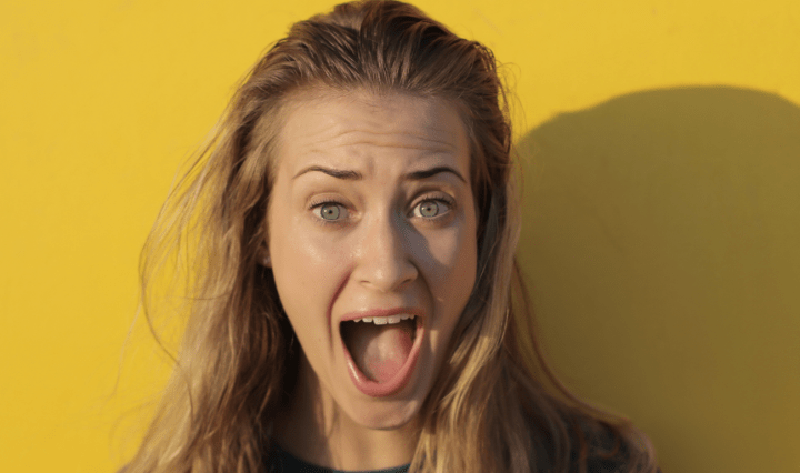 A white woman with blonde hair stands in front of a yellow war with her mouth open in shock