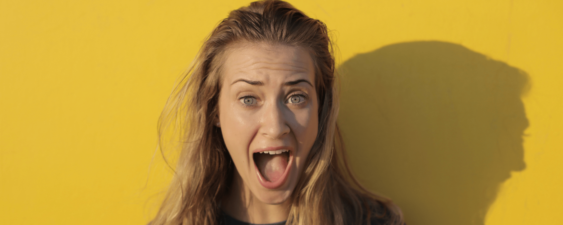 A white woman with blonde hair stands in front of a yellow war with her mouth open in shock
