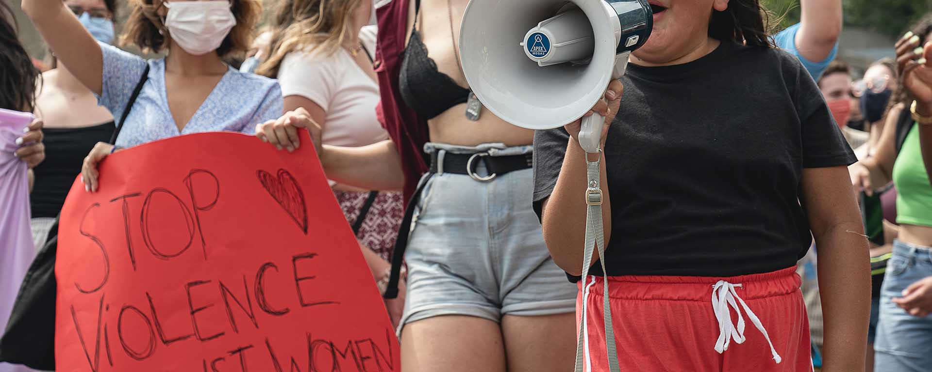 A young girl holds a megaphone at a protest. Another woman holds a sign saying "stop violence against women"