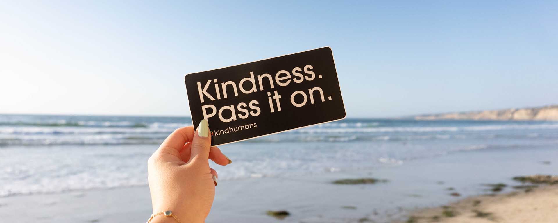 Close up of a white woman's hand. She holds a card that reads: "Kindess. Pass it on." The beach is in the background