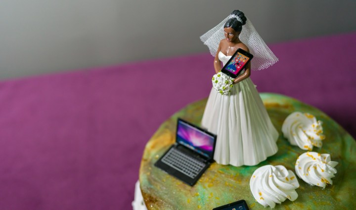 A bride cake topper on top of a wedding cake holds a mobile phone, with a laptop and ipad at her feet