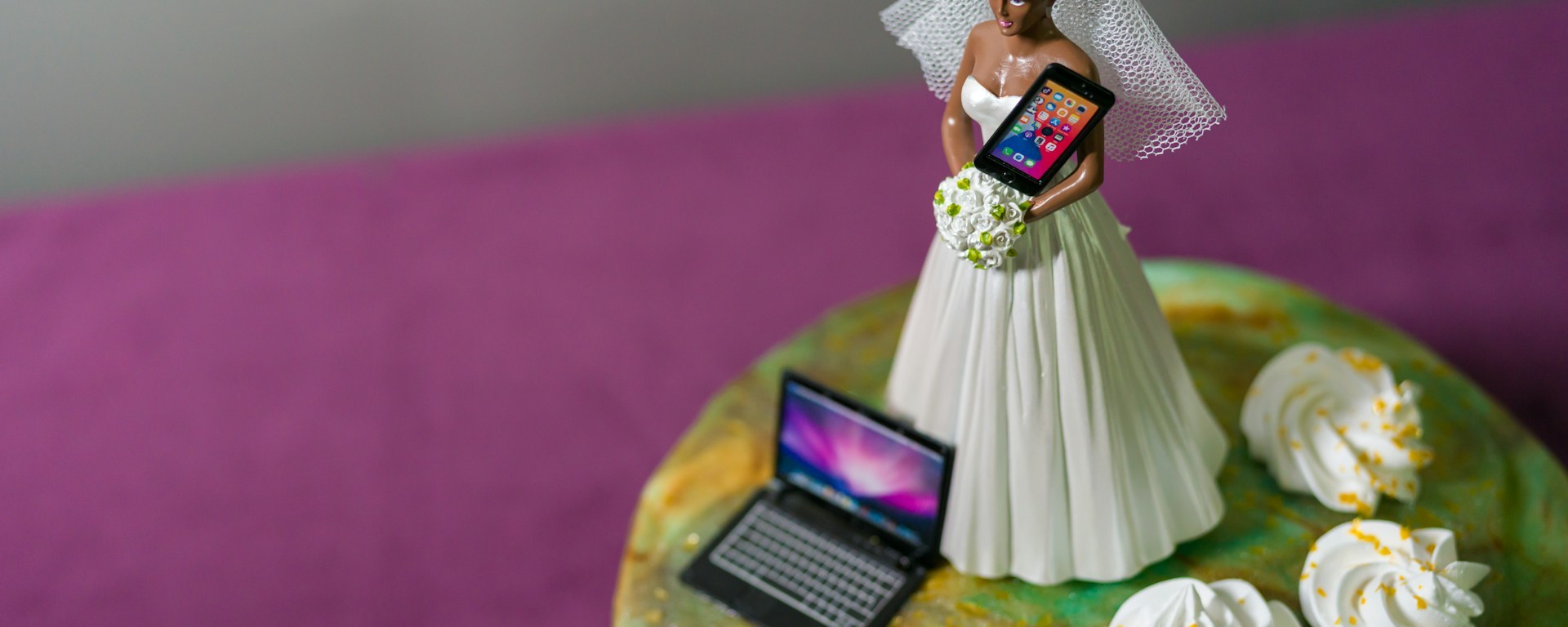A bride cake topper on top of a wedding cake holds a mobile phone, with a laptop and ipad at her feet