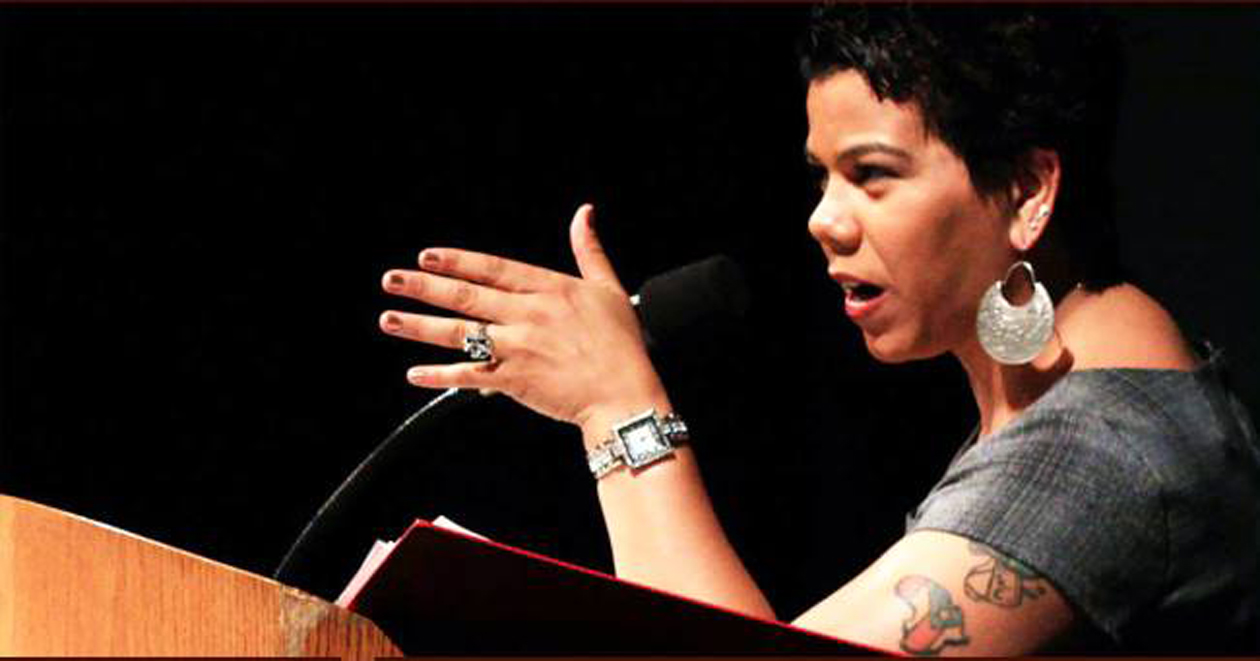 Rosa Clemente is a Black-Puerto Rican woman. She is standing at a podium pointing her hand forward as she speaks