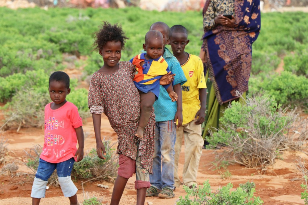 A girl holds a smaller child, with other children walking beside her and a woman in the background, in Tukaraq, Somalia