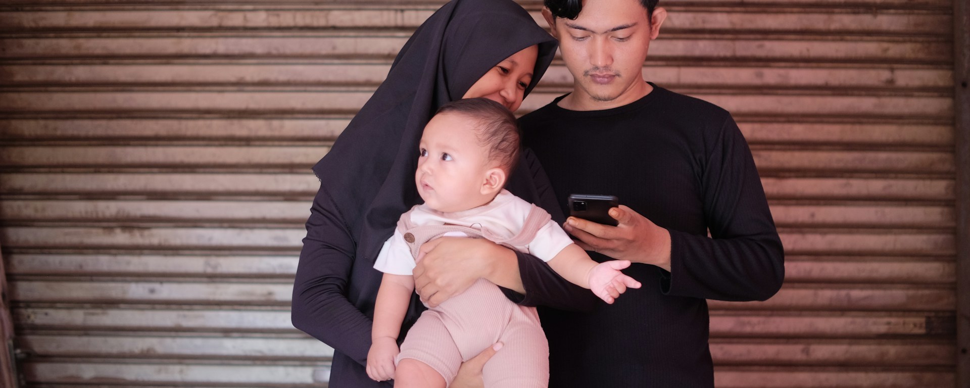 A man and woman hold a baby while looking at a phone and smile