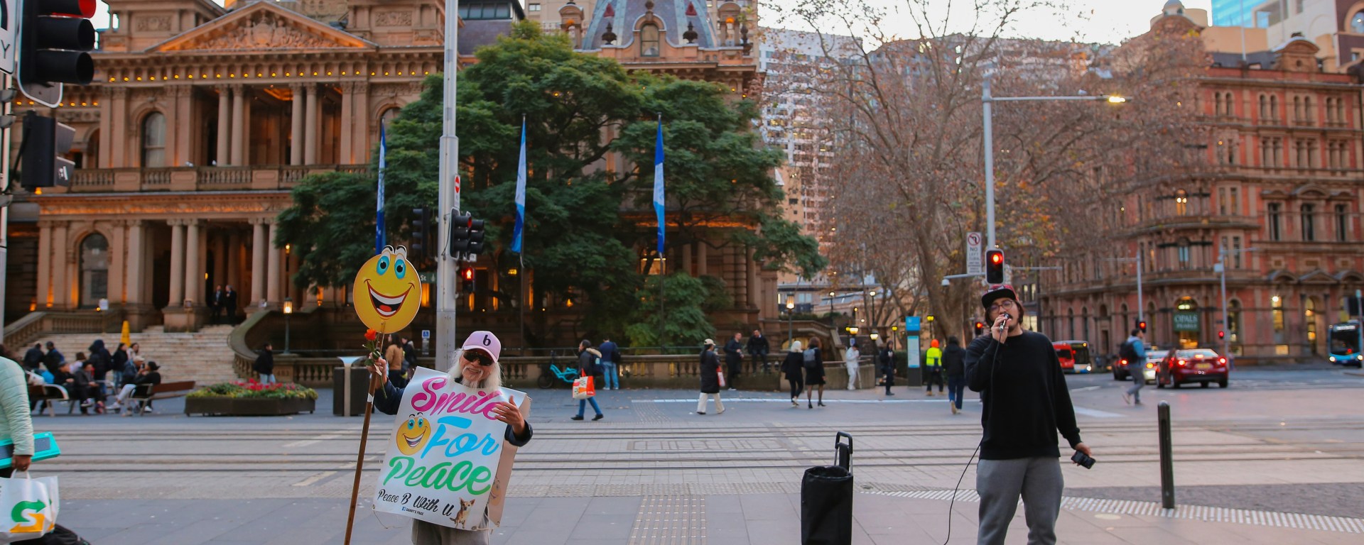 A man holds a smiling ballon and a sign that says 'smile for peace,' while another man sings on the street of Sydney