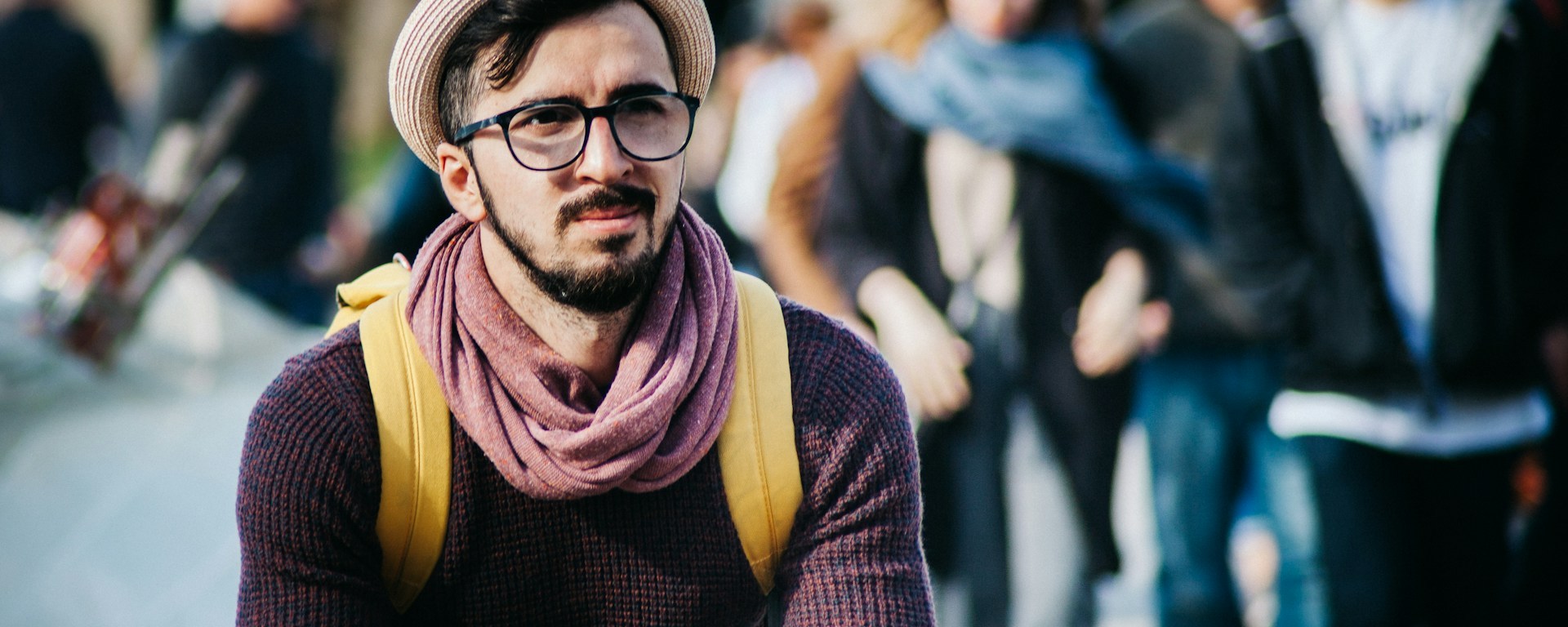 A young man is sitting down. He wears a hat and glasses, with a backpack
