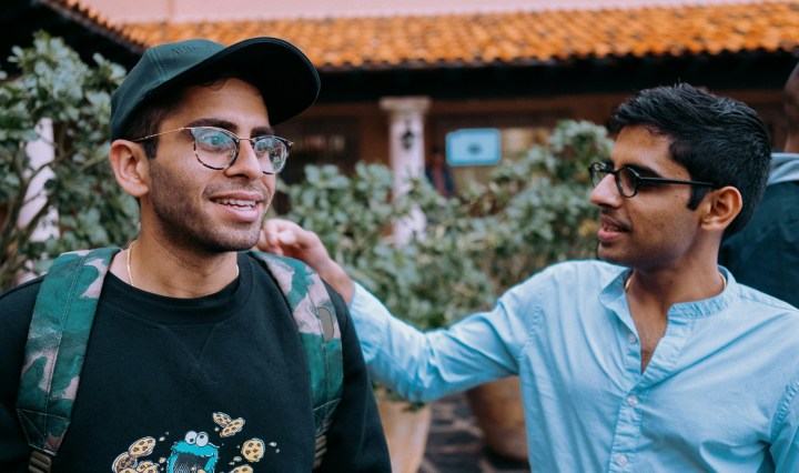 Two young men stand side by side. One of them has his hand on the other's shoulder, as he smiles