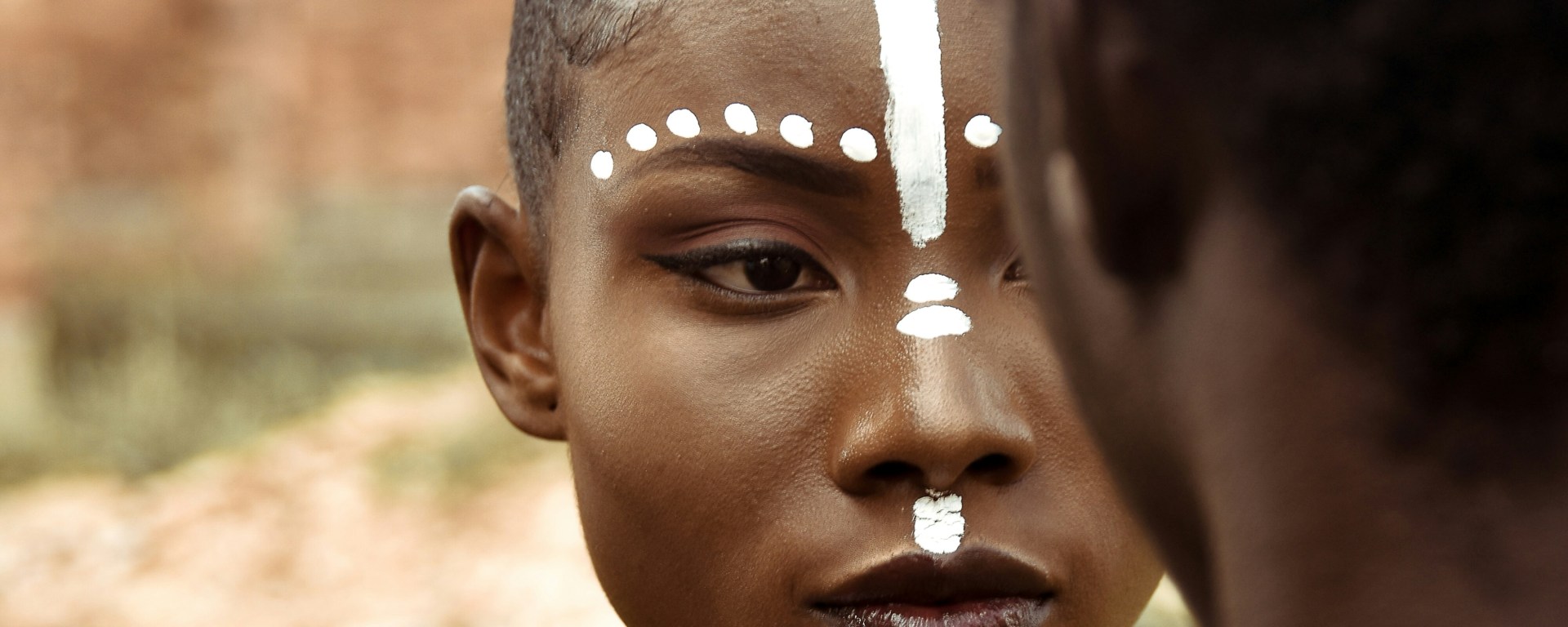 Woman from Ghana is looking into a man, who has his back to the camera. She wears white face paint in a line down her forehead, and dots around her eyebrows
