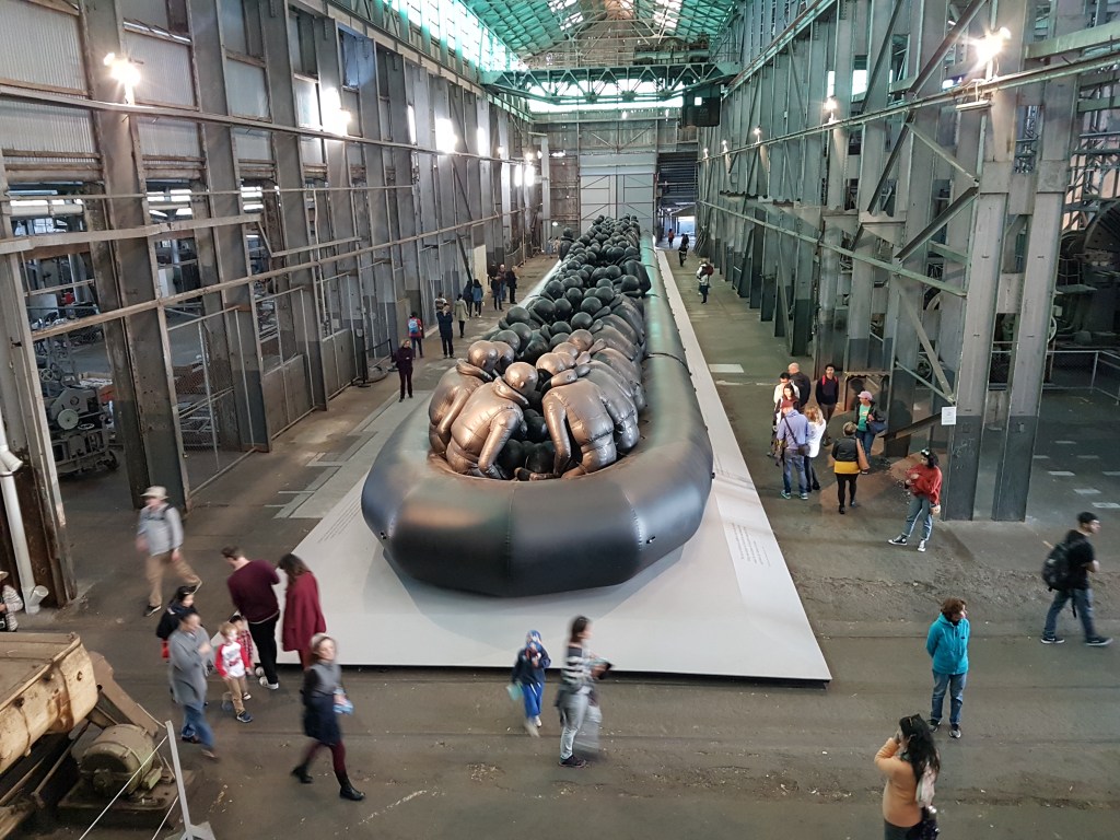 Aerial view of the installation. People walk around the raft in an industrial space, with the scupture towering over them