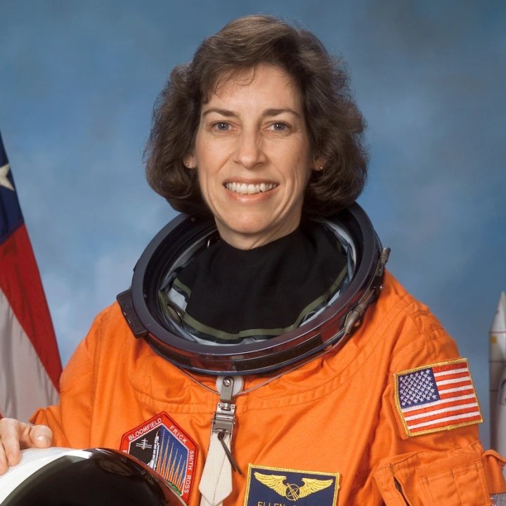 Dr Ellen Ochoa is a white Latina with wavy brown hair. She is smiling wearing an orange astronaut uniform, with the USA flag behind her