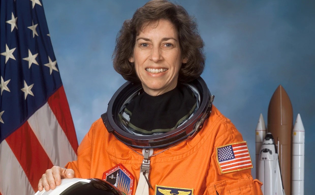 Dr Ellen Ochoa is a white Latina with wavy brown hair. She is smiling wearing an orange astronaut uniform, with the USA flag behind her