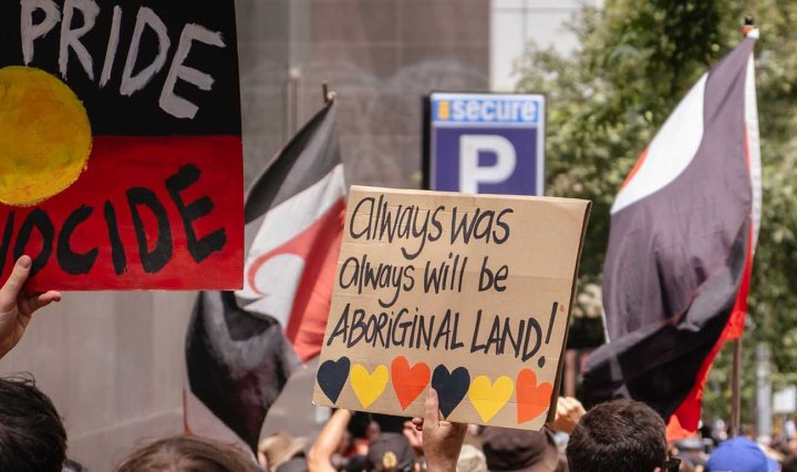 Protesters carry signs with the Aboriginal flag. One sign says: No pride in genocide. Another says: Always was always will be Aboriginal land!