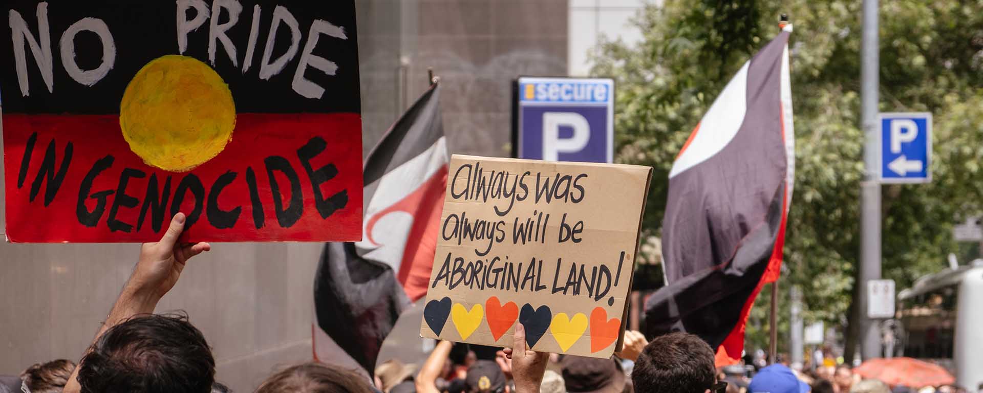 Protesters carry signs with the Aboriginal flag. One sign says: No pride in genocide. Another says: Always was always will be Aboriginal land!