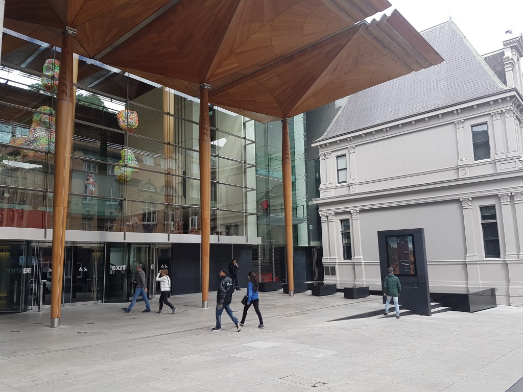 People walk outside of the Auckland Art Gallery, with impressive wooden panels above