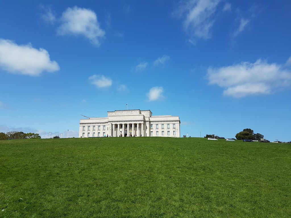 The Auckland Museum in the background, surrounded by a sprawling green lawn and a beautiful blue sky with few clouds