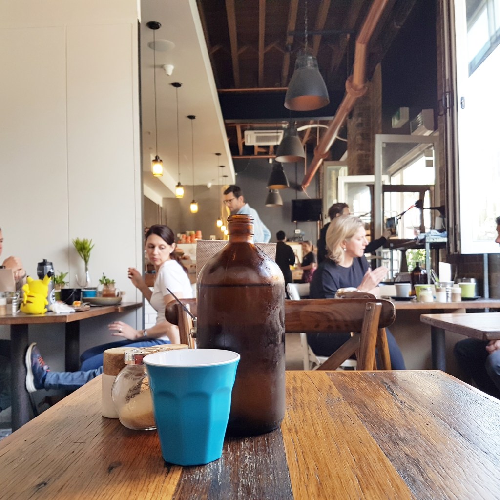 Glass and water bottle sit on a table, with people talking in a cafe in the background