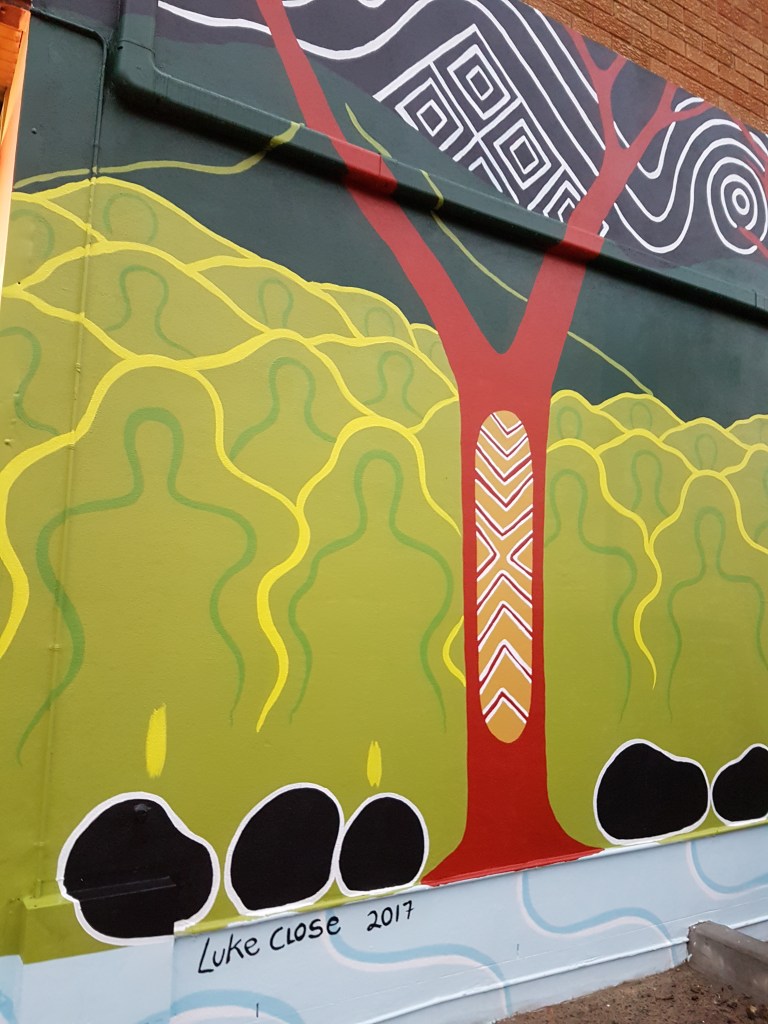 Street art outside a community organisation, showing Aboriginal art of a tree and people, signed by Luke Cose 2017