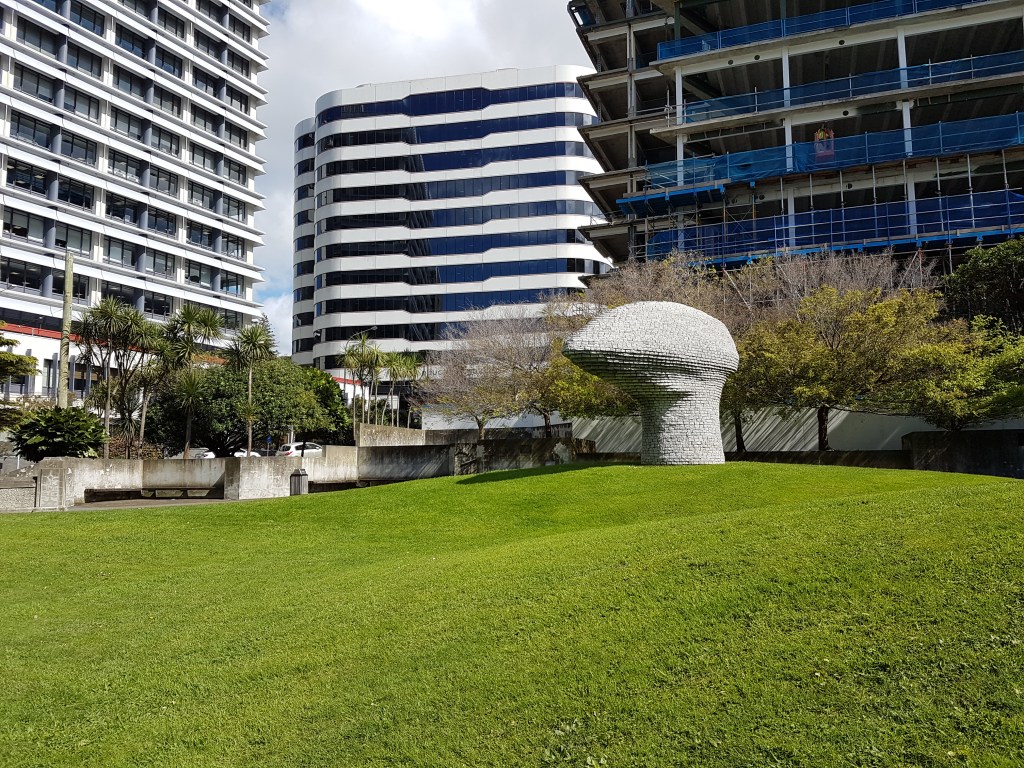 White mushroom-shaped sculpture in a garden, with buildings in the background
