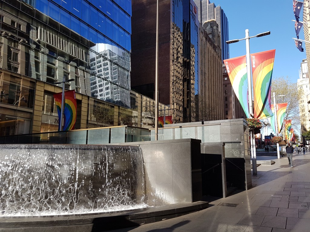 Waterfall in the Sydney CBD, with pride flags hanging from street lights, in the background 