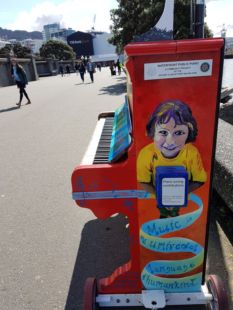 Side of a public piano outdoors is decorated with the picture of a girl and a banner saying "music"