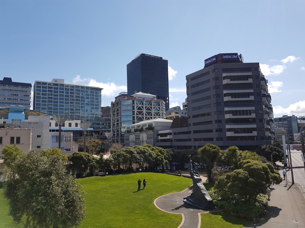 People stand in the distance of a large park with buildings in the background