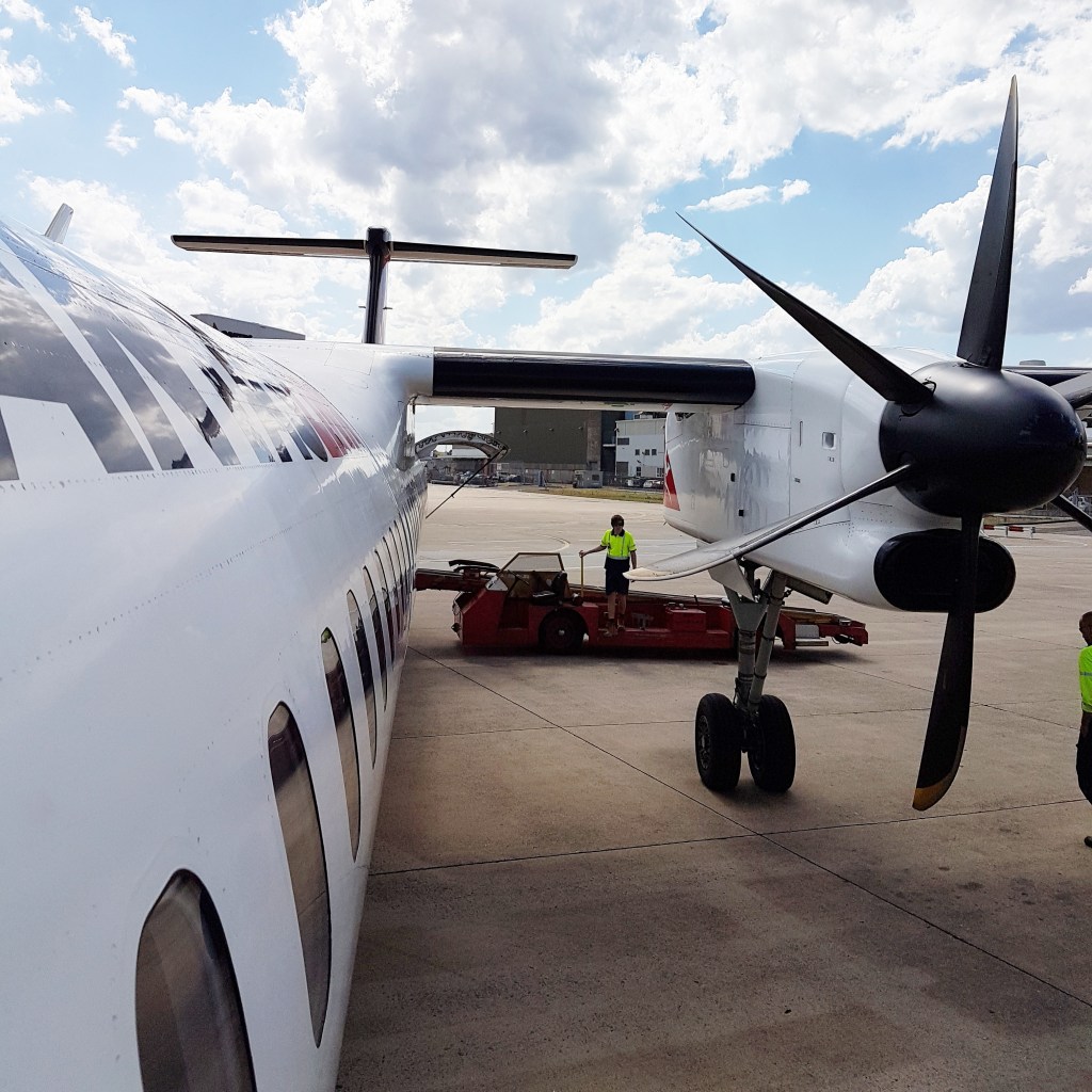 The side of a plane and its propeller . A baggage worker is seen in the background