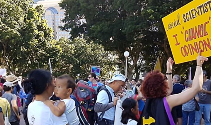Protesters in Sydney. One woman wears a cape and holds a sign that reads: Social Scientist - I change opinions