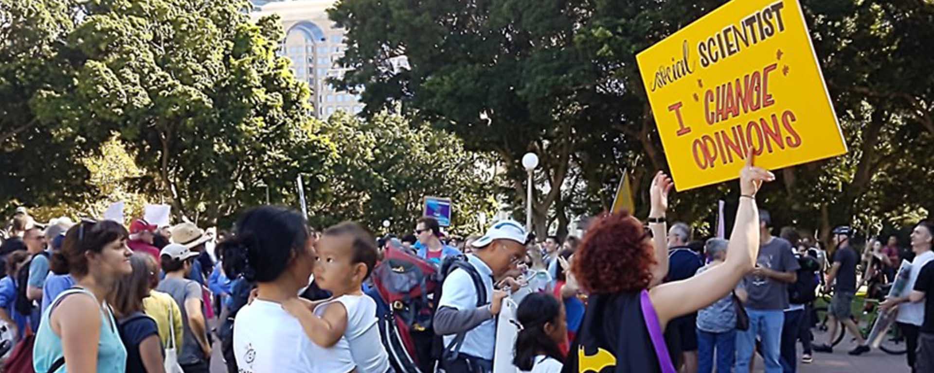 Protesters in Sydney. One woman wears a cape and holds a sign that reads: Social Scientist - I change opinions