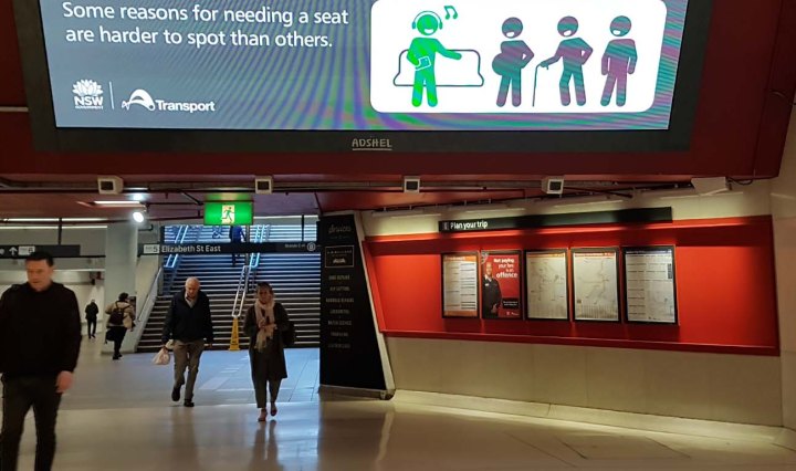 People walking in Martin Place station underground. A large monitor shows an ad with stick figure people: one is green, seated, and listening and singing to their music; the other figures are black - one is pregnant, the other uses a walking stick, and the other stands