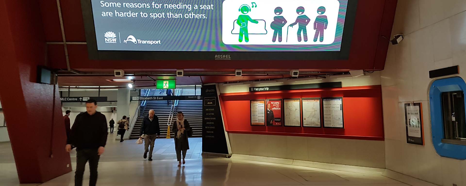 People walking in Martin Place station underground. A large monitor shows an ad with stick figure people: one is green, seated, and listening and singing to their music; the other figures are black - one is pregnant, the other uses a walking stick, and the other stands
