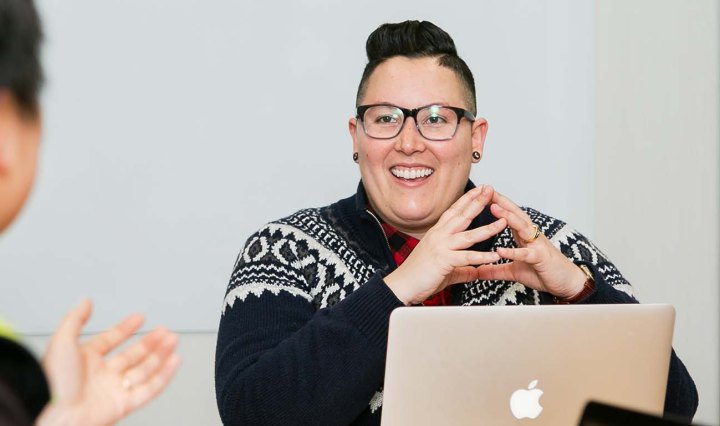 A white woman wearing glasses is smiling as another person talks to them. She has her laptop open