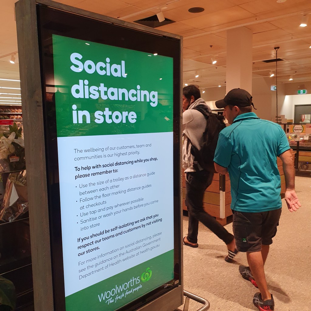 People walking into supermarket next to a dense sign from Woolworths, explaining 'social distancing in store'