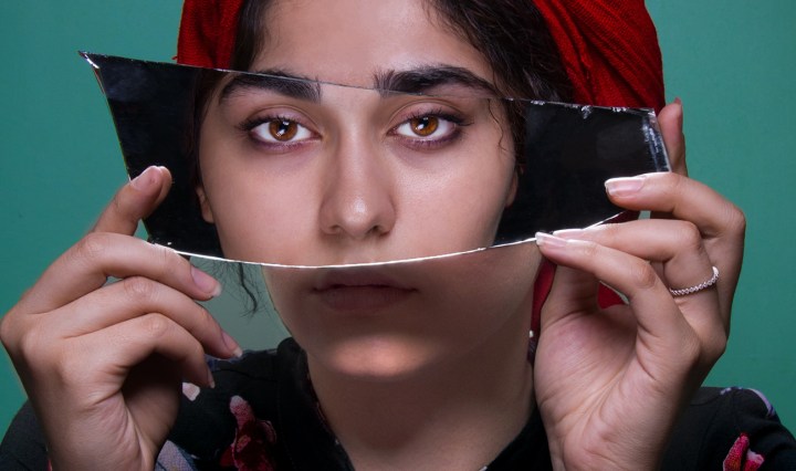 A Brown South Asian woman holds up a piece of broken mirror over her face that shows her reflection