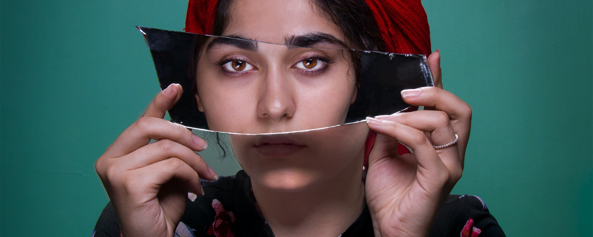 A Brown South Asian woman holds up a piece of broken mirror over her face that shows her reflection