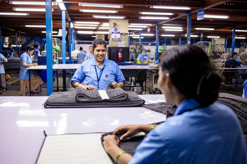 A woman in a factory is folding a mat, smiling at another younger woman who has her back to the camera