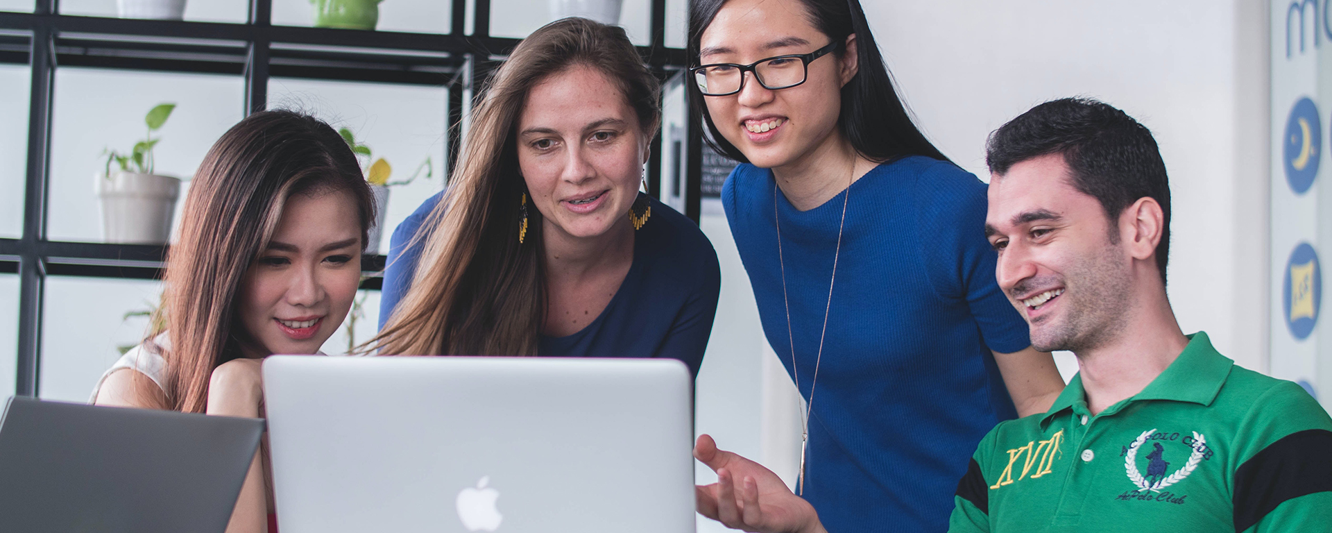 Two Asian women, one White woman, and one white man are smiling while looking at their laptop