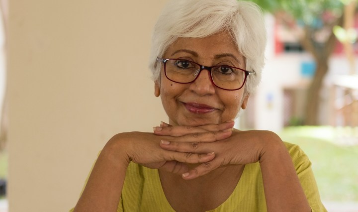 Older South Asian woman with dark skin wears glasses and smiles with her hands folded
