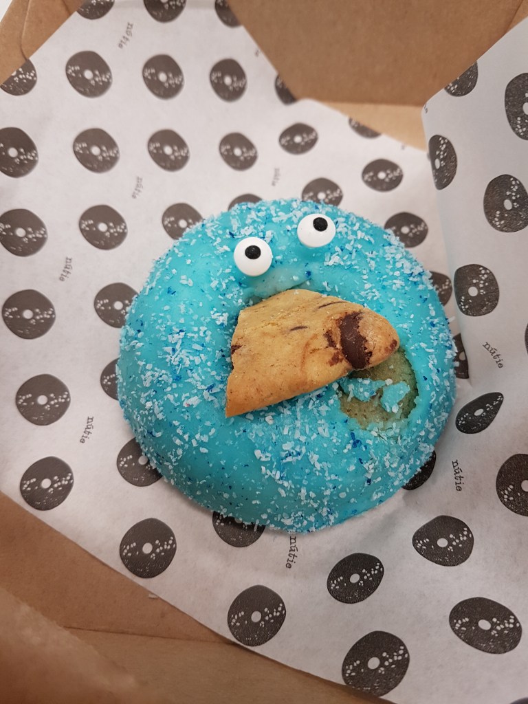 A blue icing donut with googly eyes and mouth eating a biscuit