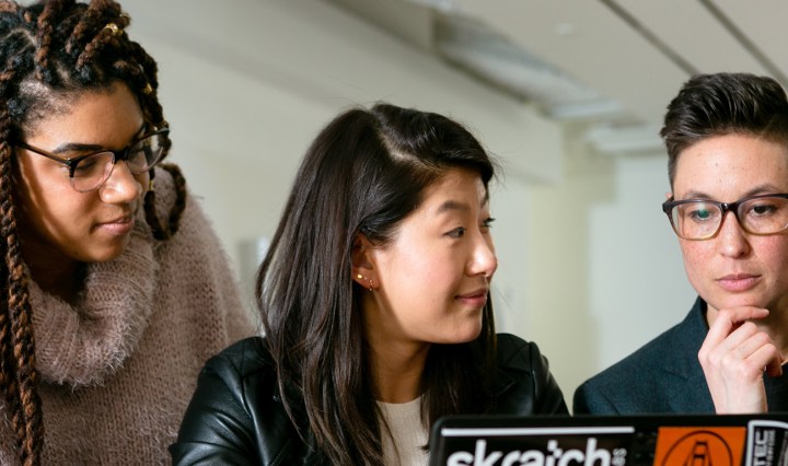 A Black woman, Asian woman, and white woman wearing the pride flag look at a laptop