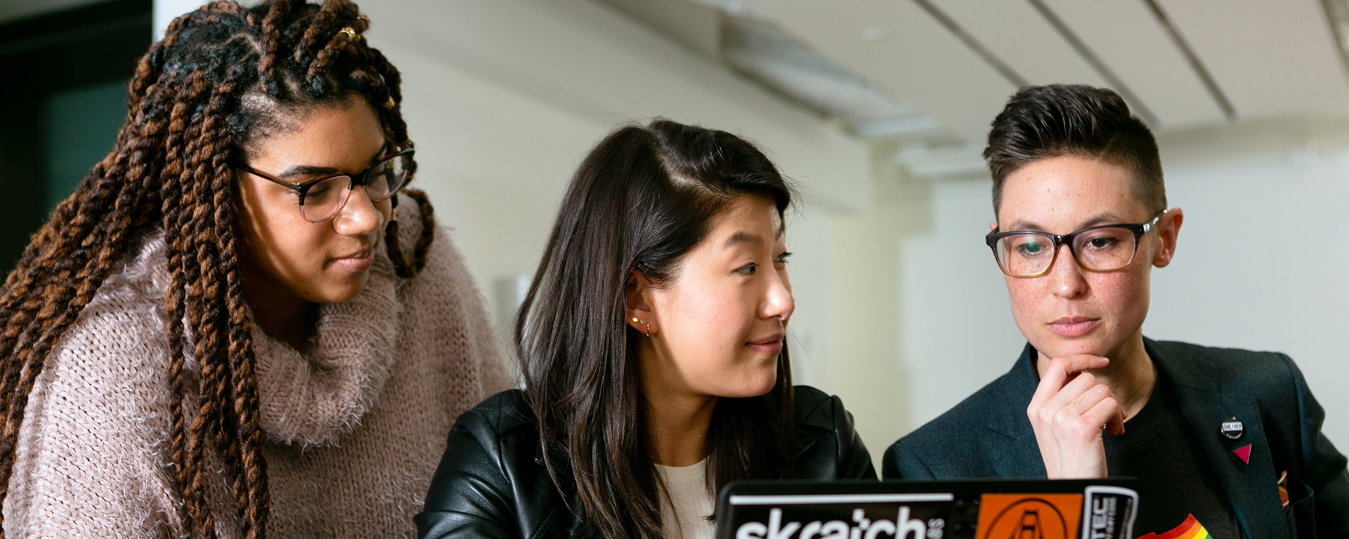 A Black woman, Asian woman, and white woman wearing the pride flag look at a laptop