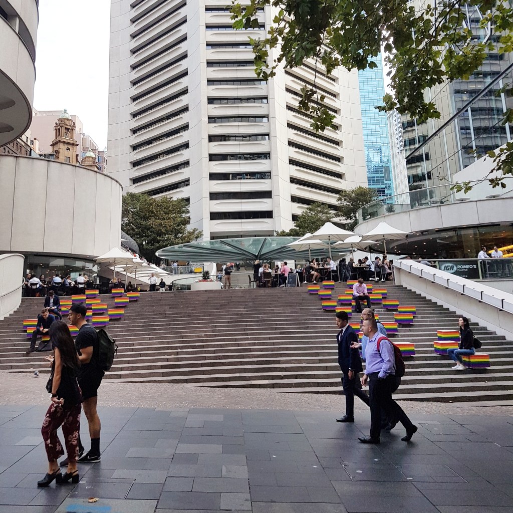 People walk across Martin Place, Sydney. In the background, people sit on stairs painted in the rainbow flag