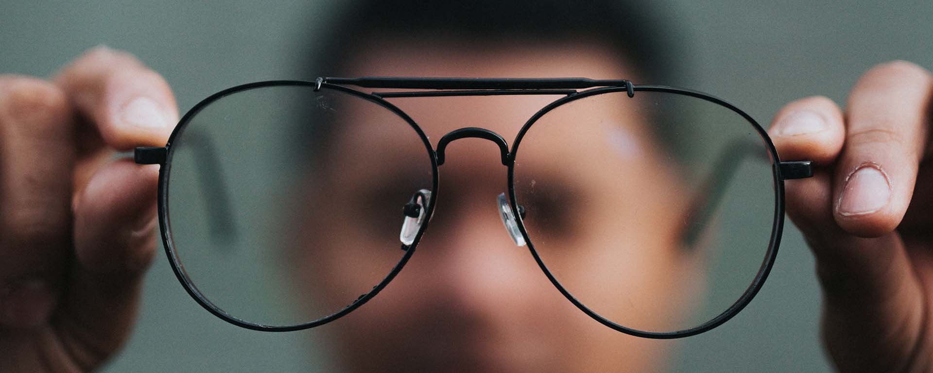 A Black man is out of focus in the background, he holds out his glasses, which are clear in the foreground