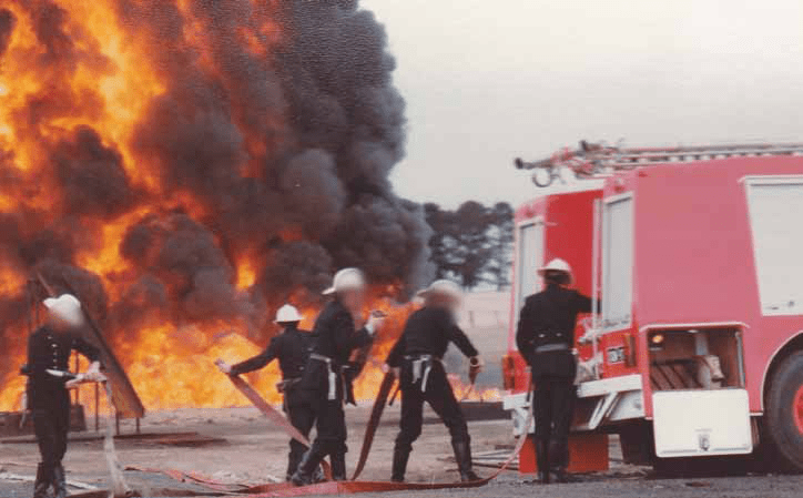 Firefighters unroll a hose next to a firetruck. A large fire blaze in the background