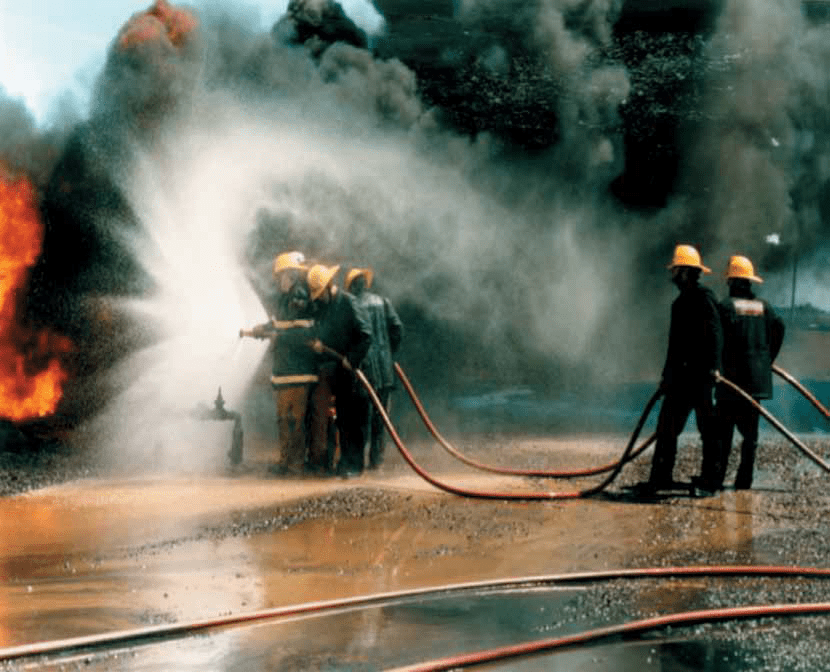 Three firefighters hold the head of the hose, while another two firefighters hold the centre of the hose. They are in front of a large fire and large black smoke