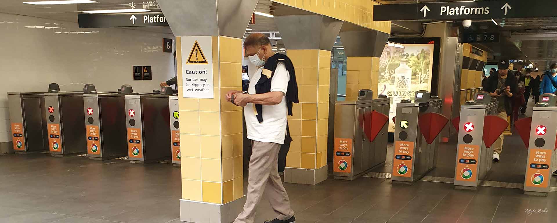 A Brown man walks across Central Station wearing a face mask. A sign behind says "Caution"