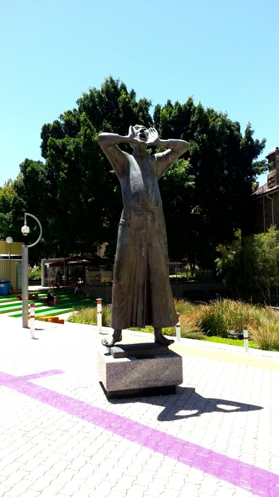 A bronze sculpture of a large person with their hands cupped around their mouth, shouting into the sky