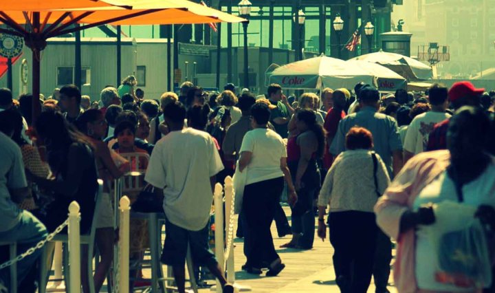 People walk in a crowded street, with some people sitting outdoors, beneath umbrellas