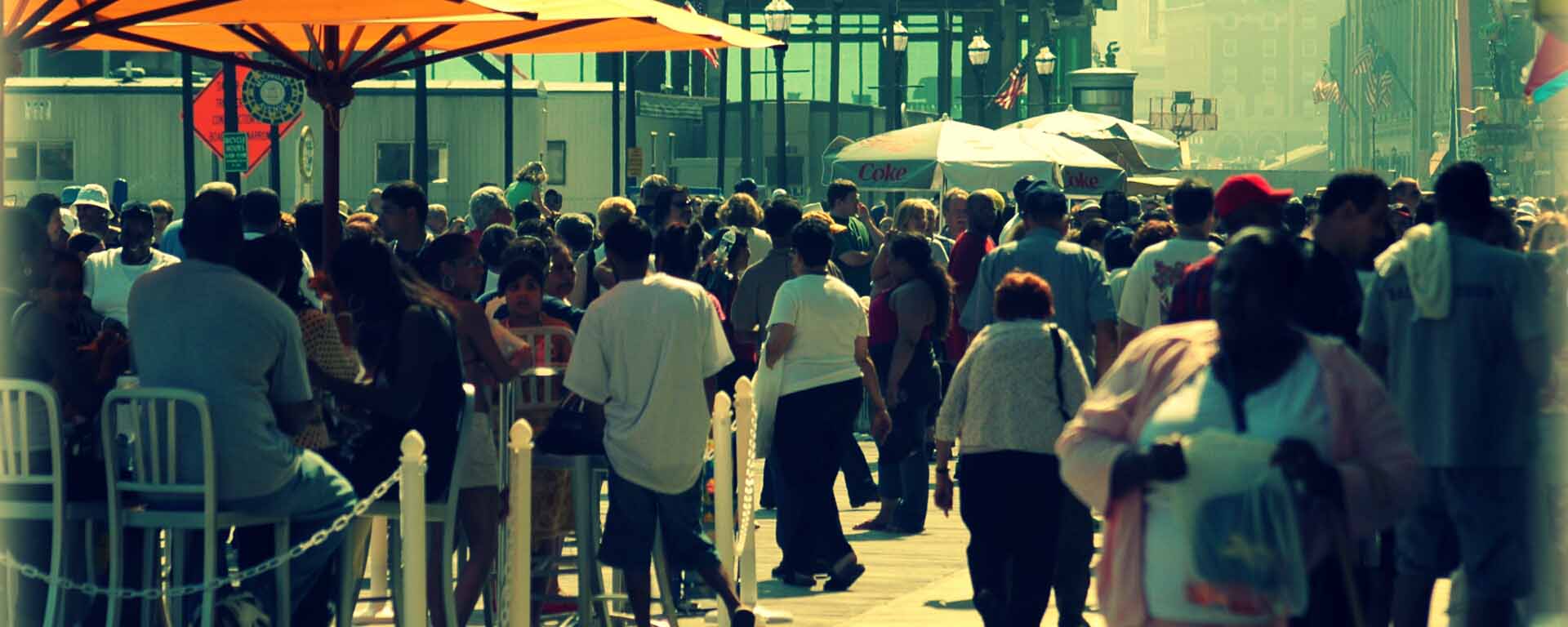 People walk in a crowded street, with some people sitting outdoors, beneath umbrellas